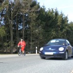 Michael Wigge mit dem Tretroller unterwegs auf einer Landstraße, neben ihm fährt ein Auto