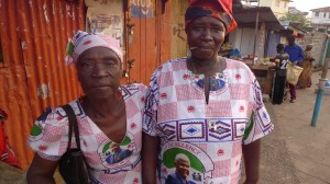 Two women wear traditional costume made out of material showing the president's face